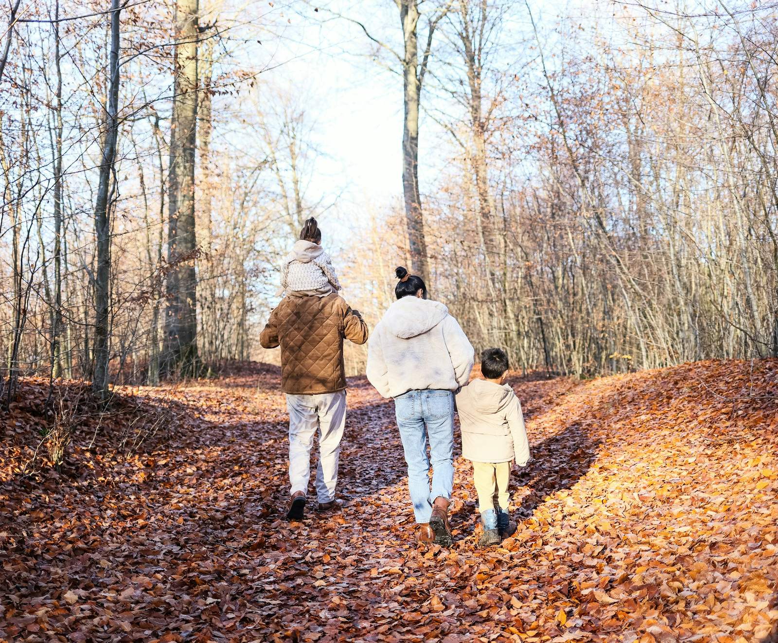 A family of four walking up a leaf-covered path through autumn woods, a child riding on the father's shoulders.
